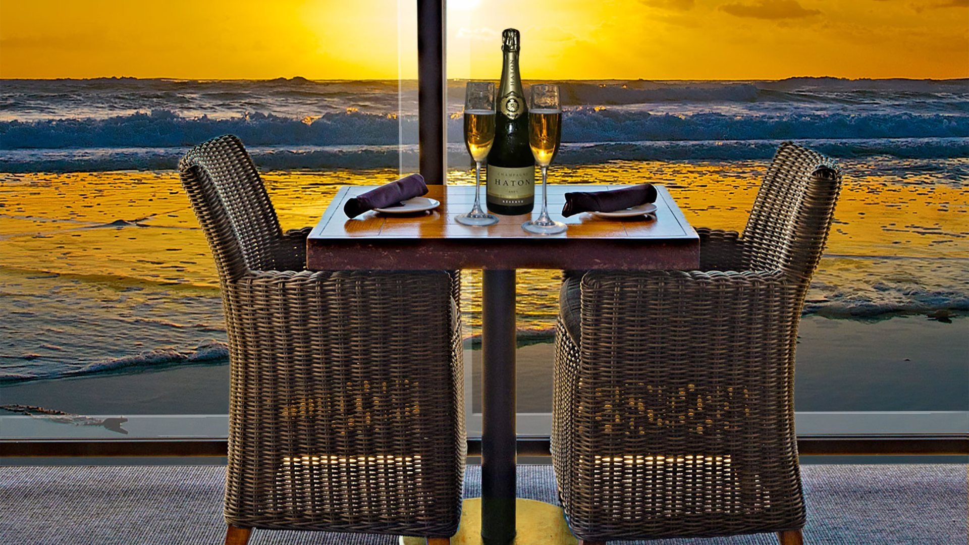 A table for two with champagne and wicker chairs facing the sunset surf at Pacific Coast Grill